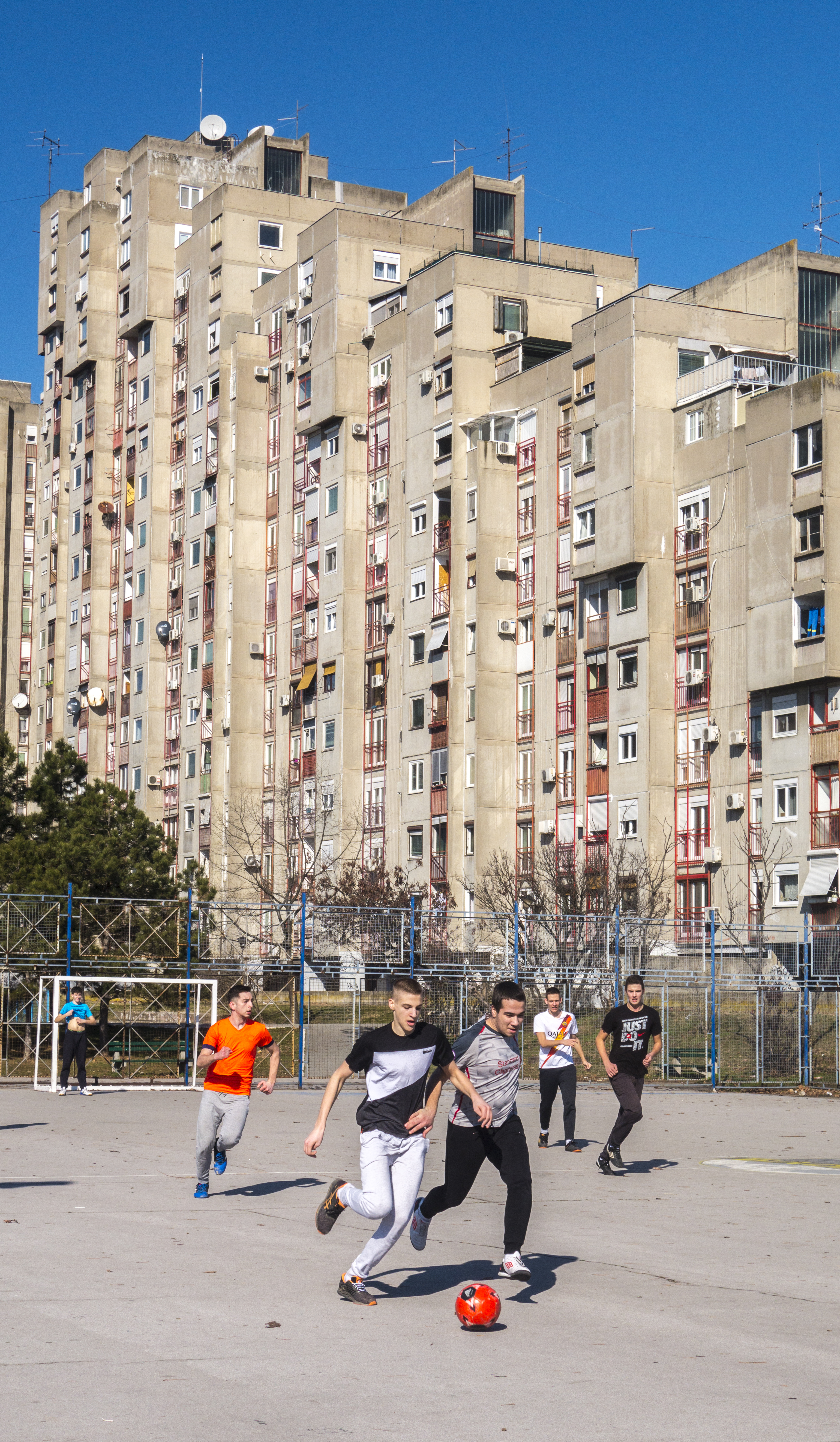 Young children play soccer on a concrete field in front of several concrete towers that belong to a mass housing project in Belgrade